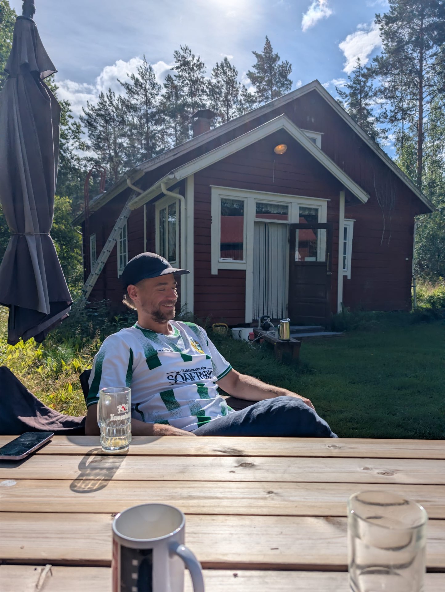 Rasmus sitter avslappnat vid ett träbord utanför en röd stuga en solig sommardag.